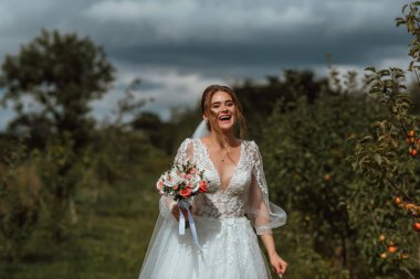 Genç ve güzel bir gelin, doğanın arka planına karşı bir düğün buketi ile poz verir; kız beyaz bir gelinlik giyer; düğün gününde bir an; mutlu kız duygularını ifade eder