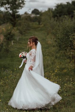 Genç ve güzel bir gelin, doğanın arka planına karşı bir düğün buketi ile poz verir; kız beyaz bir gelinlik giyer; düğün gününde bir an; mutlu kız duygularını ifade eder