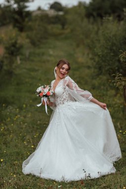 Genç ve güzel bir gelin, doğanın arka planına karşı bir düğün buketi ile poz verir; kız beyaz bir gelinlik giyer; düğün gününde bir an; mutlu kız duygularını ifade eder