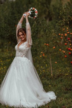 Genç ve güzel bir gelin, doğanın arka planına karşı bir düğün buketi ile poz verir; kız beyaz bir gelinlik giyer; düğün gününde bir an; mutlu kız duygularını ifade eder