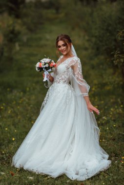 Genç ve güzel bir gelin, doğanın arka planına karşı bir düğün buketi ile poz verir; kız beyaz bir gelinlik giyer; düğün gününde bir an; mutlu kız duygularını ifade eder