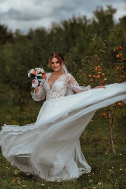 Genç ve güzel bir gelin, doğanın arka planına karşı bir düğün buketi ile poz verir; kız beyaz bir gelinlik giyer; düğün gününde bir an; mutlu kız duygularını ifade eder