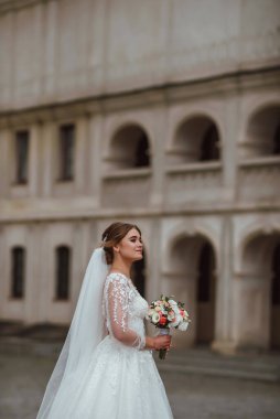 Genç ve güzel bir gelin, doğanın arka planına karşı bir düğün buketi ile poz verir; kız beyaz bir gelinlik giyer; düğün gününde bir an; mutlu kız duygularını ifade eder