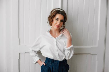 Beyaz gömlekli ve kot pantolonlu seksi bir kız beyaz bir kapının önünde poz veriyor.;