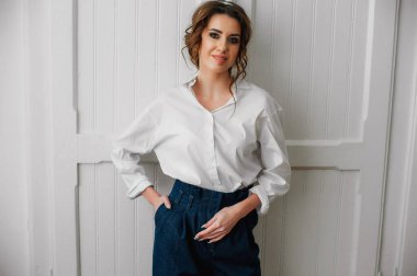 Beyaz gömlekli ve kot pantolonlu seksi bir kız beyaz bir kapının önünde poz veriyor.;