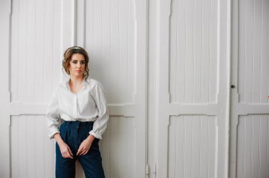 Beyaz gömlekli ve kot pantolonlu seksi bir kız beyaz bir kapının önünde poz veriyor.;