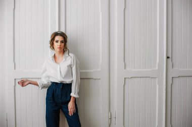 Beyaz gömlekli ve kot pantolonlu seksi bir kız beyaz bir kapının önünde poz veriyor.;