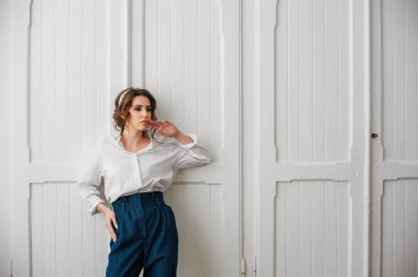 Beyaz gömlekli ve kot pantolonlu seksi bir kız beyaz bir kapının önünde poz veriyor.;