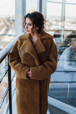 Uzun kürk giyen seksi bir kız balkonda poz veriyor. Kış şehrinin arka planında.;