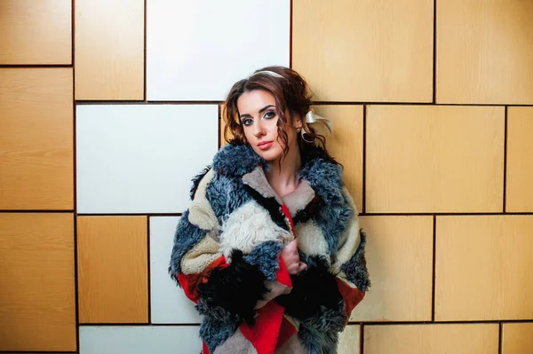 a sexy girl dressed in a long fur coat poses indoors against the wall;