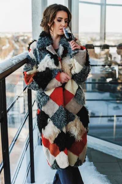 a sexy girl dressed in a long fur coat poses on the balcony against the background of the winter city; holding a glass of red wine