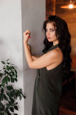 Kıvırcık, seksi esmer, kafeteryada poz veriyor; koyu renk bir elbise giyiyor; dudakları kırmızı rujla boyanmış.