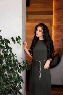 Kıvırcık, seksi esmer, kafeteryada poz veriyor; koyu renk bir elbise giyiyor; dudakları kırmızı rujla boyanmış.