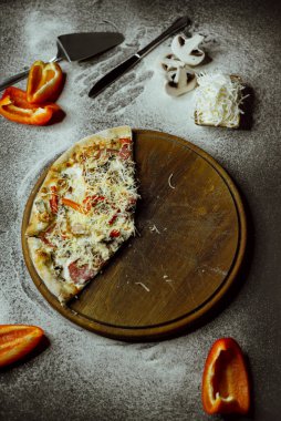 on a black background there is a pizza on a board, scattered flour, pepper, mushrooms and utensils; sliced pizza; not a whole pizza on the board; pieces of pizza on the table