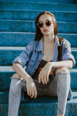 a teenage girl with long dark hair poses for the camera; sits in a spectator seat at the stadium; a model-looking girl on the street; slender figure of a beautiful girl in a bikini under denim clothes; wearing sunglasses