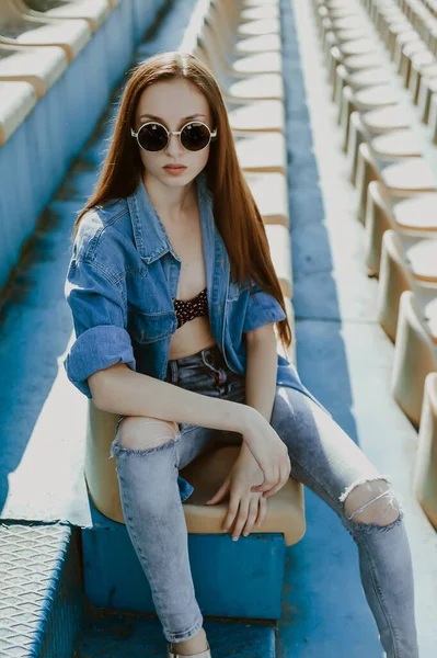 a teenage girl with long dark hair poses for the camera; sits in a spectator seat at the stadium; a model-looking girl on the street; slender figure of a beautiful girl in a bikini under denim clothes; wearing sunglasses