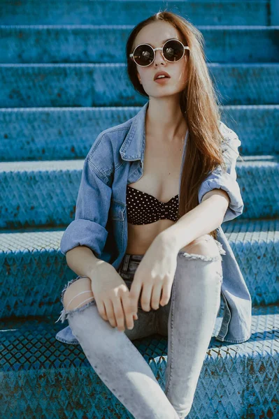 a teenage girl with long dark hair poses for the camera; sits in a spectator seat at the stadium; a model-looking girl on the street; slender figure of a beautiful girl in a bikini under denim clothes; wearing sunglasses