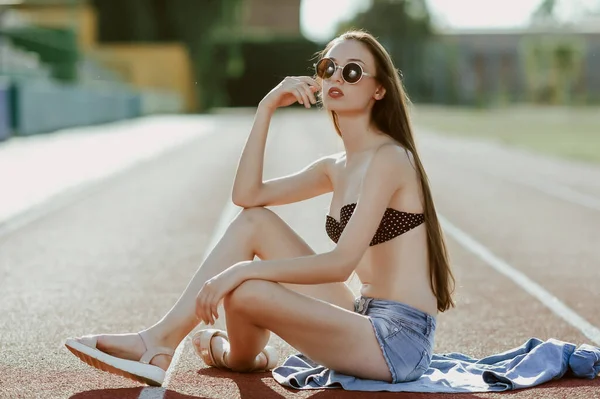a teenage girl with long dark hair poses for the camera; on a stadium running track; a model-looking girl on the street; slender figure of a beautiful girl in a bikini and denim shorts; sunglasses on his head