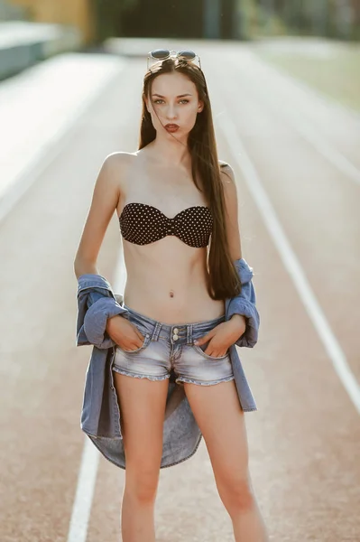 a teenage girl with long dark hair poses for the camera; on a stadium running track; a model-looking girl on the street; slender figure of a beautiful girl in a bikini and denim shorts; sunglasses on his head