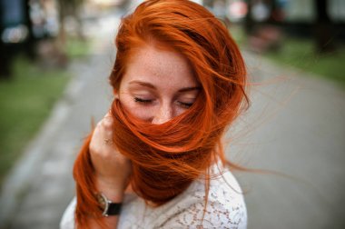 a red-haired girl in a light skirt; a very cheerful girl with long red hair; lips painted with red lipstick; sits on a bench and has a good mood; poses for a photographer; street photo session 