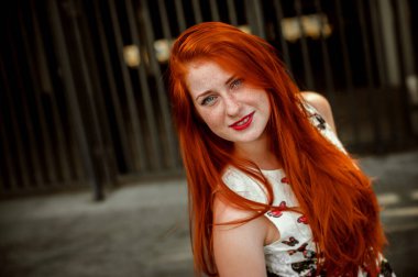 a red-haired girl in a light skirt; a very cheerful girl with long red hair; lips painted with red lipstick; sits on a bench and has a good mood; poses for a photographer; street photo session 