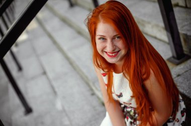 a red-haired girl in a light skirt; a very cheerful girl with long red hair; lips painted with red lipstick; sits on a bench and has a good mood; poses for a photographer; street photo session 