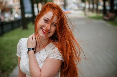 a red-haired girl in a light skirt; a very cheerful girl with long red hair; lips painted with red lipstick; sits on a bench and has a good mood; poses for a photographer; street photo session 