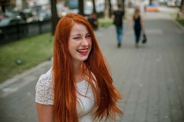 a red-haired girl in a light skirt; a very cheerful girl with long red hair; lips painted with red lipstick; sits on a bench and has a good mood; poses for a photographer; street photo session