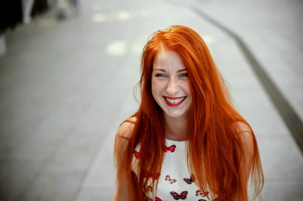 a red-haired girl in a light skirt; a very cheerful girl with long red hair; lips painted with red lipstick; sits on a bench and has a good mood; poses for a photographer; street photo session