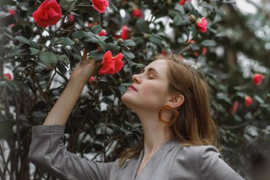 A girl poses in a botanical garden among plants. The model is wearing a gray suit. Freckles on the girl's face. The girl poses among flowers, bushes, trees, cacti.