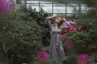A girl poses in a botanical garden among plants. The model is wearing a gray suit. Freckles on the girl's face. The girl poses among flowers, bushes, trees, cacti.
