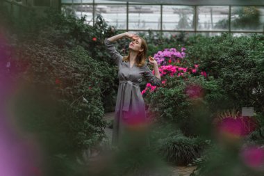 A girl poses in a botanical garden among plants. The model is wearing a gray suit. Freckles on the girl's face. The girl poses among flowers, bushes, trees, cacti.