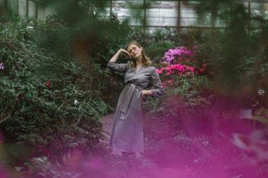 A girl poses in a botanical garden among plants. The model is wearing a gray suit. Freckles on the girl's face. The girl poses among flowers, bushes, trees, cacti.