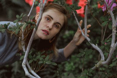 A girl poses in a botanical garden among plants. The model is wearing a gray suit. Freckles on the girl's face. The girl poses among flowers, bushes, trees, cacti.