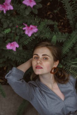 A girl poses in a botanical garden among plants. The model is wearing a gray suit. Freckles on the girl's face. The girl poses among flowers, bushes, trees, cacti.