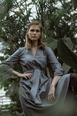 A girl poses in a botanical garden among plants. The model is wearing a gray suit. Freckles on the girl's face. The girl poses among flowers, bushes, trees, cacti.