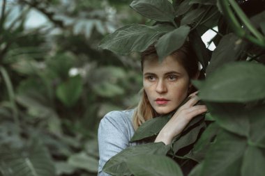A girl poses in a botanical garden among plants. The model is wearing a gray suit. Freckles on the girl's face. The girl poses among flowers, bushes, trees, cacti.