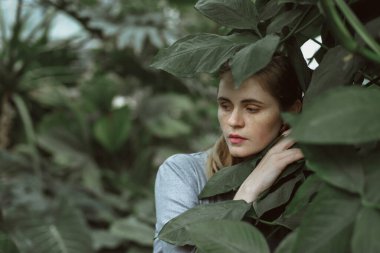 A girl poses in a botanical garden among plants. The model is wearing a gray suit. Freckles on the girl's face. The girl poses among flowers, bushes, trees, cacti.