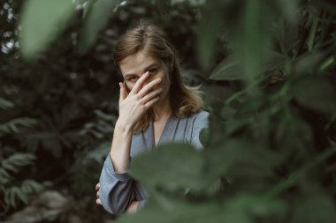 A girl poses in a botanical garden among plants. The model is wearing a gray suit. Freckles on the girl's face. The girl poses among flowers, bushes, trees, cacti.