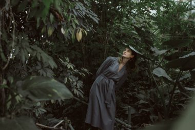 A girl poses in a botanical garden among plants. The model is wearing a gray suit. Freckles on the girl's face. The girl poses among flowers, bushes, trees, cacti.