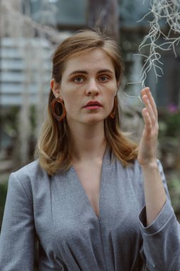 A girl poses in a botanical garden among plants. The model is wearing a gray suit. Freckles on the girl's face. The girl poses among flowers, bushes, trees, cacti.