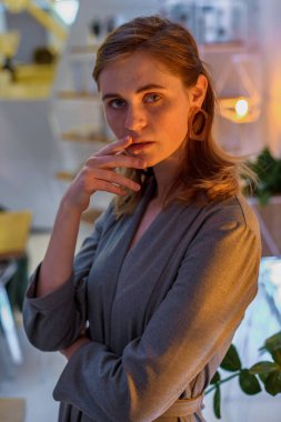 The girl poses in a cafe. The model is wearing a gray suit. Freckles on the girl's face. A fair-haired girl poses in a lit room near a lamp