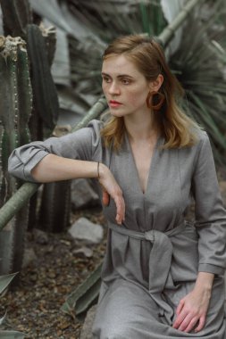 A girl poses in a botanical garden among plants. The model is wearing a gray suit. Freckles on the girl's face. The girl poses among flowers, bushes, trees, cacti.
