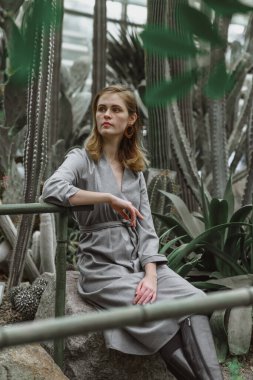 A girl poses in a botanical garden among plants. The model is wearing a gray suit. Freckles on the girl's face. The girl poses among flowers, bushes, trees, cacti.
