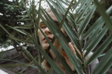 A girl poses in a botanical garden among plants. The model is wearing a gray suit. Freckles on the girl's face. The girl poses among flowers, bushes, trees, cacti.