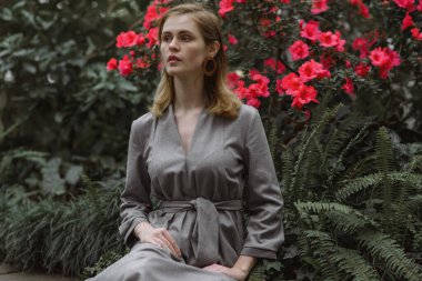 A girl poses in a botanical garden among plants. The model is wearing a gray suit. Freckles on the girl's face. The girl poses among flowers, bushes, trees, cacti.