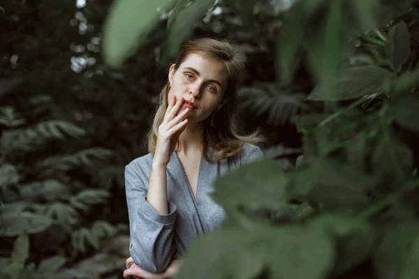 A girl poses in a botanical garden among plants. The model is wearing a gray suit. Freckles on the girl's face. The girl poses among flowers, bushes, trees, cacti.