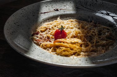 Dishes from the chef of the restaurant. A dish on a table in a cafe. Appetizing food from the restaurant menu. Fresh and tasty food. Carbonara in a plate with a small tomato under a streak of sunlight