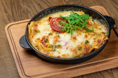 Dishes from the chef of the restaurant. A dish on a table in a cafe. Appetizing food from the restaurant menu. Fresh and tasty food. Lasagna sprinkled with greens and tomatoes in a pan on a board
