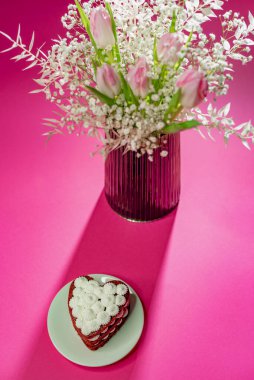 Dessert in the form of a heart in a plate near flowers on a pink background. white delicate flowers in a vase. A light dessert with several layers.